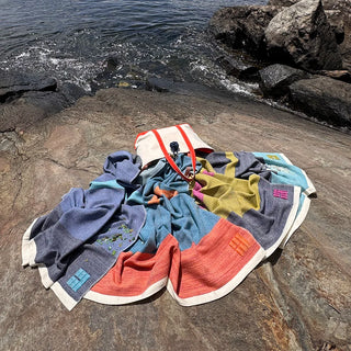 A group of multicolor handwoven wool throws with white trim and a canvas tote back with orange handles is laid out on a rocky shoreline next to the ocean water.