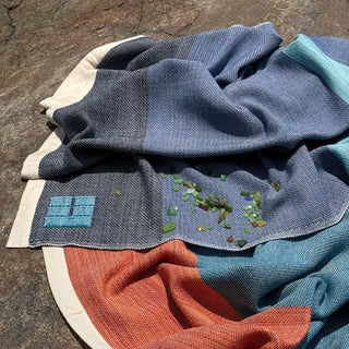 A folded Swans Island handwoven throw in blue, teal, and rust-orange sections with a cream border, placed on a rough stone surface. Small green and brown bits of sea glass are scattered on top, and an aqua weaver's mark is visible on one corner.