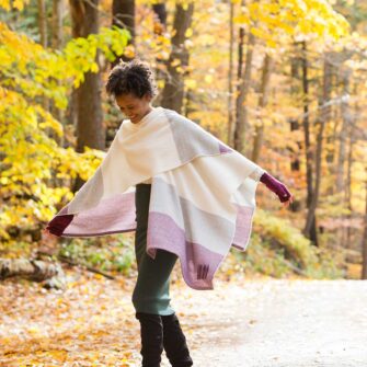 Swans Island Company's Maine Coast Cape. Handwoven in Maine with soft organic merino wool and touch of alpaca. Colors are dyed with all natural dyes. Shown here in Beetroot on woman walking with yellow fall foliage in the background.