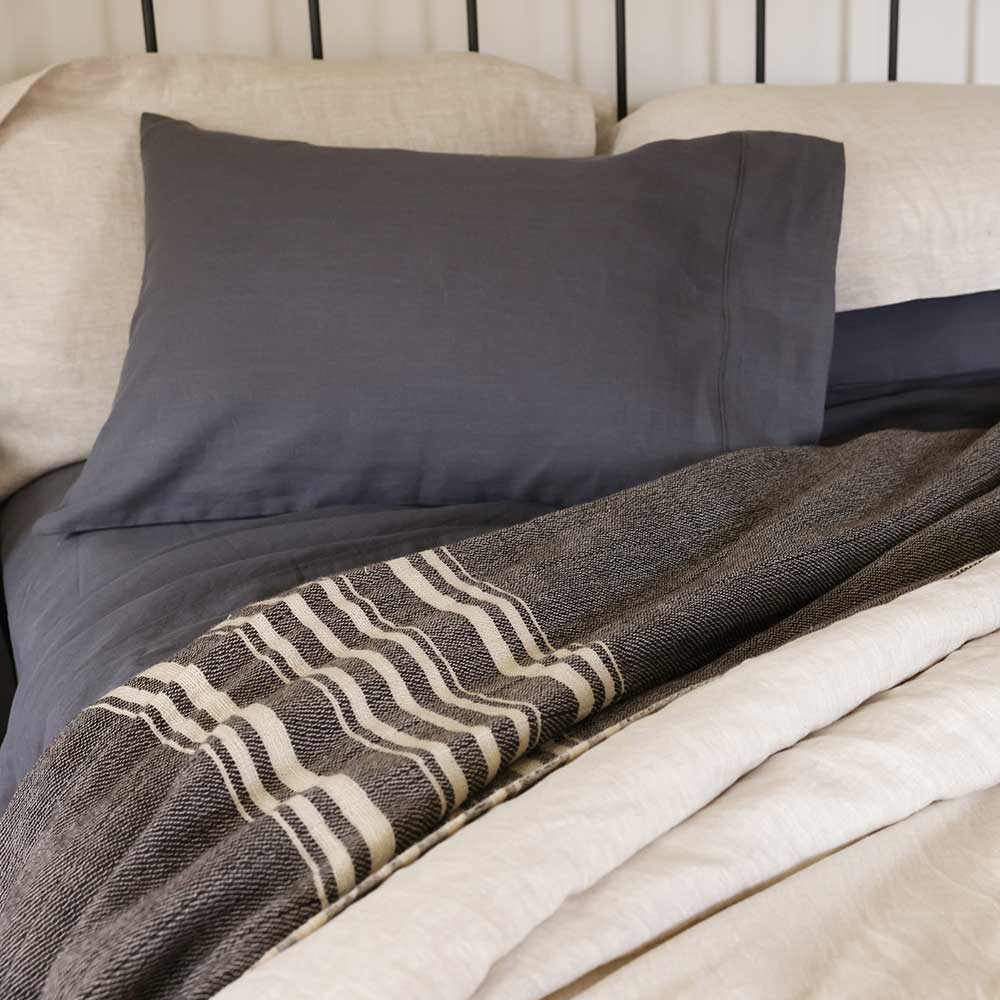 Swans Island Company's handwoven Grace Winter Blanket. Shown here in Charcoal with natural stripes. Handwoven in Maine with hand-dyed locally sourced wool yarns. Shown with Relaxed Linen Sheet set in Obsidian.