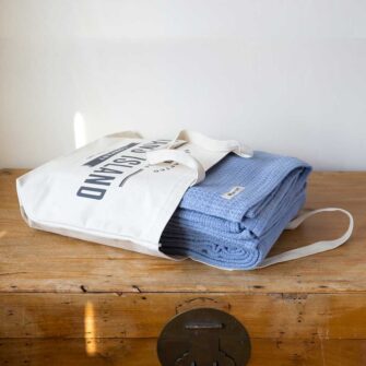 Swans Island Company's Tuscany Cotton Blanket. Soft 100% Cotton blanket woven with an allover basketweave texture. Made in USA with undyed natural colored yarn. Shown here in French Blue. Each blankets comes in our signature cotton canvas tote.