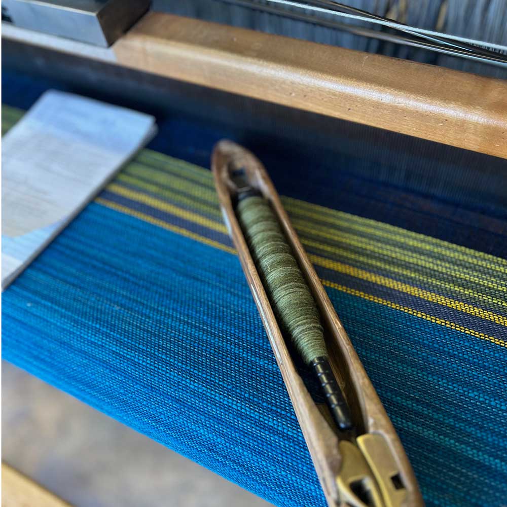 The Swans Island & Eric Hopkins Ltd. Edition Throw 2024. A shuttle with hand-dyed green wool is ready to be woven at the Swans Island studio in Northport, Maine. On the loom at the Swans Island studio in Northport, Maine.