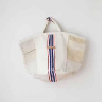 Swans Island Company's Quilt Basket Bag by Govou. Handcrafted in Spain from vintage linen grain sacks. Long canvas shoulder straps and short handles, lined with cotton. Each bag is unique. Shown here in natural with red and blue stripes.