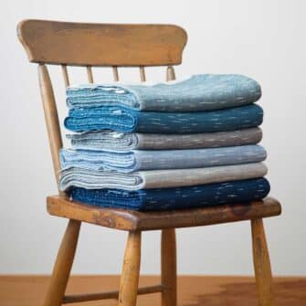 Swans Island Company Firefly Throw. Stack in Blue Spruce, Sapphire Blue, Teal, Stonewall Grey, Sea Salt Grey and Twilight Blue. 100% soft merino wool, handwoven in Maine.
