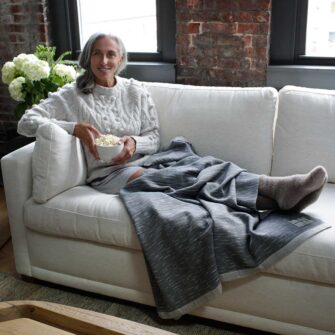 Swans Island Company Firefly Throw in Stonewall dark grey, draped on woman eating popcorn on a couch. 100% soft merino wool, handwoven in Maine.