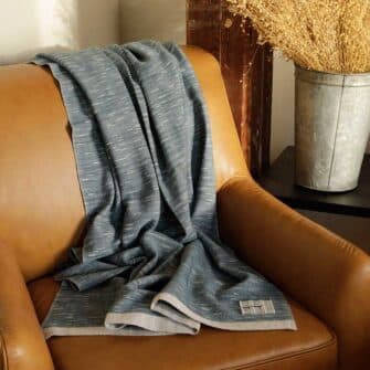 Swans Island Company Firefly Throw in Stonewall dark grey draped on a leather chair. 100% soft merino wool, handwoven in Maine.