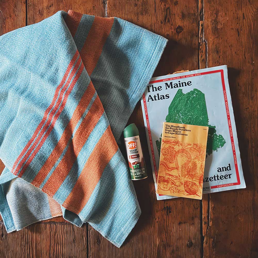 Swans Island x Pierrepont Hicks Wool Camp Blanket - handwoven in Maine with hand-dyed wool. Inspired by camp blankets of the past, woven in colors of today - a bold turquoise ground with shades of orange stripes in the borders. Blanket folded on a vintage wide pine plank floor with Maine road atlas and other camp accoutrements.