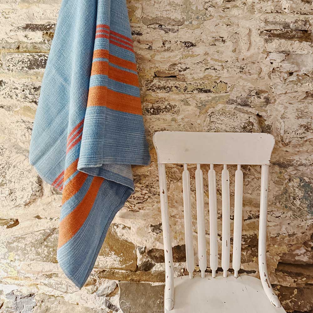 Swans Island x Pierrepont Hicks Wool Camp Blanket - handwoven in Maine with hand-dyed wool. Inspired by camp blankets of the past, woven in colors of today - a bold turquoise ground with shades of orange stripes in the borders. Blanket draped on a vintage white painted chair against a whitewashed, rustic stone camp wall.
