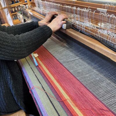 Swans Island Company's 2025 Limited Edition "Autumn in New England" Throw commemorates Yankee Magazines' 90th anniversary. This handwoven throw features 90 color changes in vibrant autumn hues of hand-dyed color. Made in Maine. Swans Island weaver at the loom making a throw.