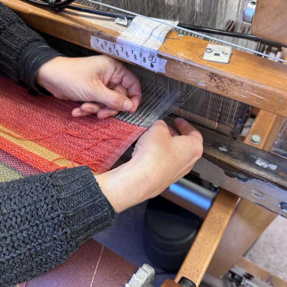 Swans Island Company's 2025 Limited Edition "Autumn in New England" Throw commemorates Yankee Magazines' 90th anniversary. This handwoven throw features 90 color changes in vibrant autumn hues of hand-dyed color. Made in Maine. Closeup detail of Swans Island weaver at the loom making a throw.