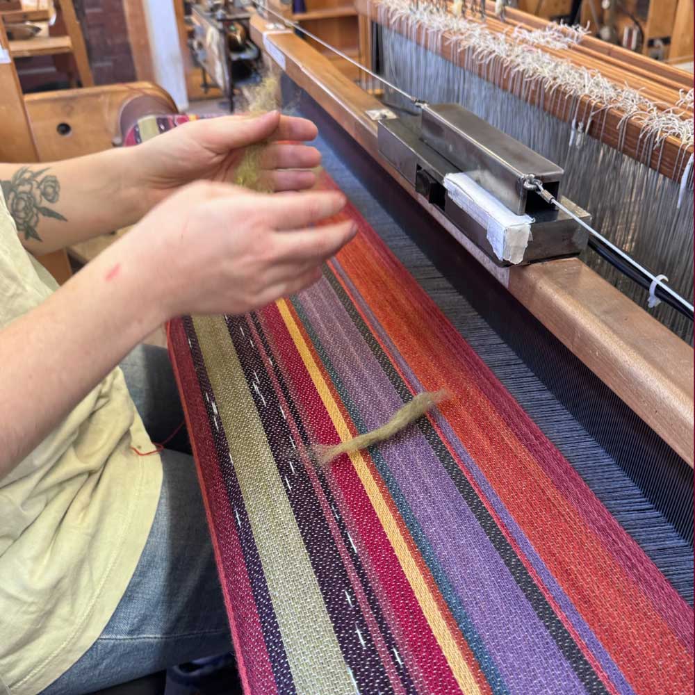 Swans Island Company's 2025 Limited Edition "Autumn in New England" Throw commemorates Yankee Magazines' 90th anniversary. This handwoven throw features 90 color changes in vibrant autumn hues of hand-dyed color. Made in Maine. Closeup detail of Swans Island weaver at the loom adding roving inlays to a throw.
